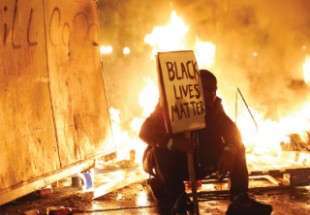 متظاهر يرفع لافتة كتب عليها «أرواح السود مهمة» وخلفه نيران المواجهات في فيرجسون بولاية ميسوري